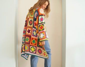 May include: A long, colorful crochet cardigan with a patchwork design featuring stars. The cardigan is made with a variety of colors, including red, orange, yellow, green, blue, and purple. The model is wearing blue jeans and white sneakers.