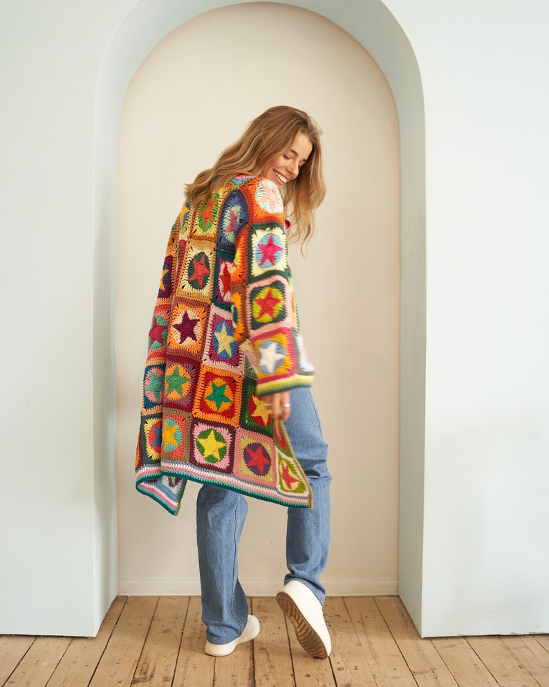 Op de afbeelding: Een lange, kleurrijke gehaakte cardigan met een patchwork-ontwerp met sterren. De cardigan is gemaakt met verschillende kleuren, waaronder rood, oranje, geel, groen, blauw en paars. Het model draagt ​​een blauwe jeans en witte sneakers.