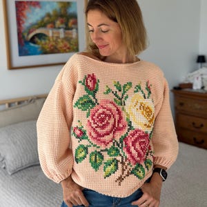 May include: A peach-colored long-sleeve sweater featuring a prominent cross-stitch floral pattern on the front. The design displays large pink and yellow roses with green leaves and stems. The sweater has a wide neckline and cuffed sleeves.