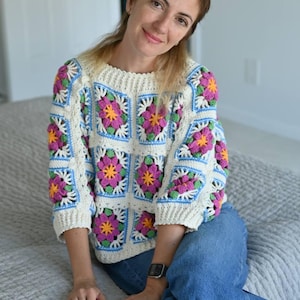 May include: A cream-coloured crochet sweater with a floral granny square design in pink, yellow, and blue. The sweater has a round neckline and three-quarter sleeves. The person is wearing blue jeans.