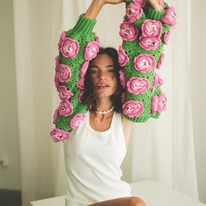 May include: A woman wearing a green and pink crocheted sweater with large flower embellishments. She is wearing a white tank top underneath and has her arms raised above her head.