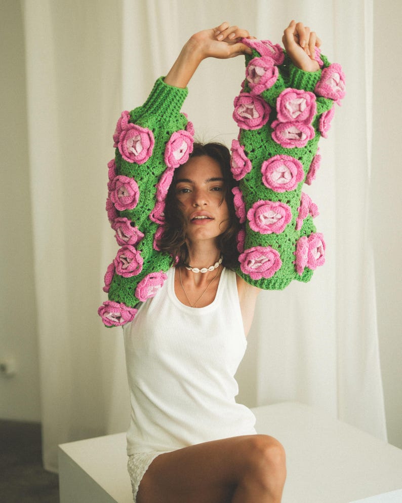 May include: A woman wearing a green and pink crocheted sweater with large flower embellishments. She is wearing a white tank top underneath and has her arms raised above her head.