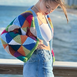 May include: A woman wearing a colorful crocheted cardigan with a white tank top and blue jeans.