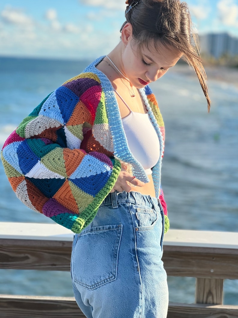 May include: A woman wearing a colorful crocheted cardigan with a white tank top and blue jeans.