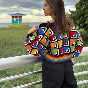 May include: A woman wearing a colorful crocheted cropped sweater with a rainbow trim. The sweater features a geometric pattern of squares and triangles in various colors, including red, orange, yellow, green, blue, purple, and black. The woman is standing on a wooden walkway with a railing.