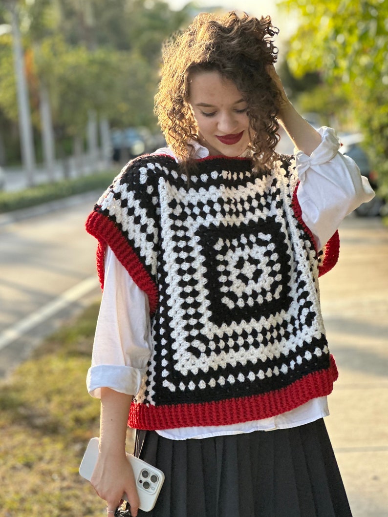 May include: A black and white crochet vest with a red trim. The vest has a square pattern with a smaller square in the center.