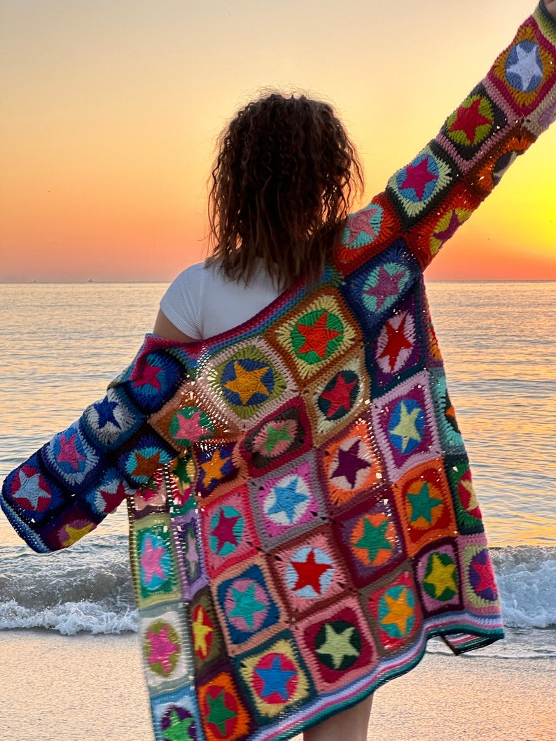 Op de afbeelding: Een kleurrijk gebreid vest met een sterpatroon. Het vest is gemaakt van verschillende gekleurde garens en heeft een losse pasvorm. Het vest wordt gedragen door een persoon die op een strand staat.