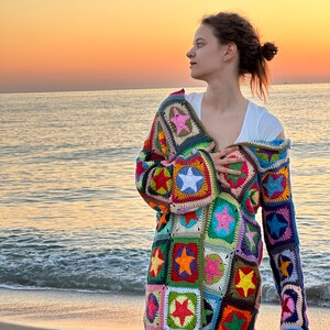 Op de afbeelding: Een vrouw draagt een kleurrijk gebreid vest met een sterpatroon. Het vest is gemaakt van verschillende gekleurde garens en heeft een lange, losse pasvorm. De vrouw staat op een strand met de zee op de achtergrond.
