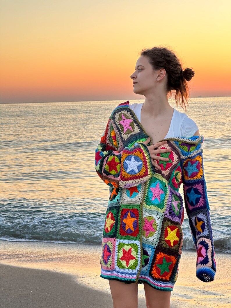 Op de afbeelding: Een vrouw draagt een kleurrijk gebreid vest met een sterpatroon. Het vest is gemaakt van verschillende gekleurde garens en heeft een lange, losse pasvorm. De vrouw staat op een strand met de zee op de achtergrond.
