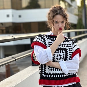 May include: A woman wearing a black and white crochet vest with red trim, a white button-down shirt, and a black skirt. She is standing in front of a building with a railing.