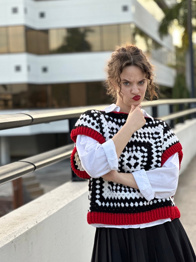 May include: A woman wearing a black and white crochet vest with red trim, a white button-down shirt, and a black skirt. She is standing in front of a building with a railing.