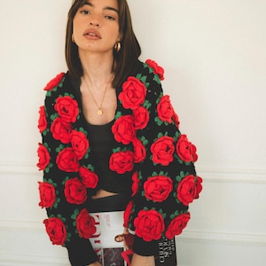 May include: A black crocheted cardigan with red roses. The cardigan is cropped and has long sleeves. The model is wearing a black top and black pants.