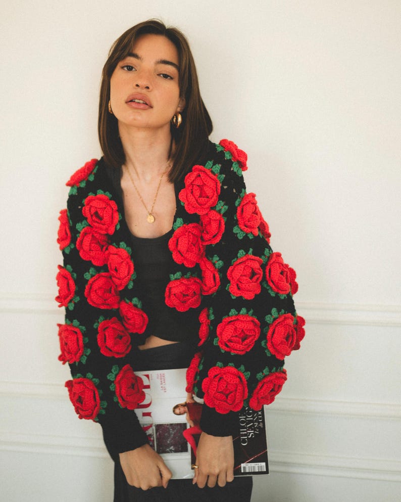 May include: A black crocheted cardigan with red roses. The cardigan is cropped and has long sleeves. The model is wearing a black top and black pants.