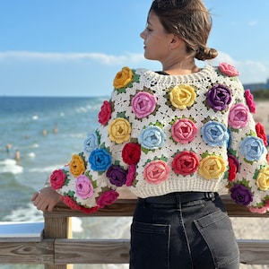 May include: A white crochet top with colorful flower embellishments. The top is cropped and has a relaxed fit. The model is wearing black jeans and is standing on a wooden pier.