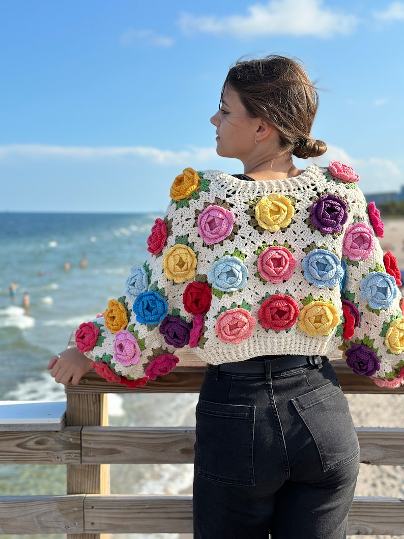 May include: A white crochet top with colorful flower embellishments. The top is cropped and has a relaxed fit. The model is wearing black jeans and is standing on a wooden pier.