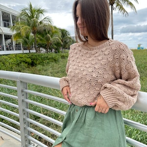 May include: A woman wearing a beige crocheted sweater with a green linen skirt. The sweater has a textured design with raised bumps.