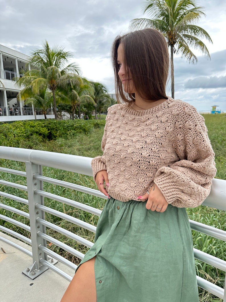 May include: A woman wearing a beige crocheted sweater with a green linen skirt. The sweater has a textured design with raised bumps.