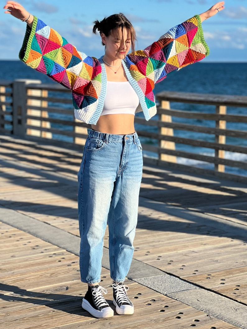 May include: A woman wearing a colorful crocheted cardigan with a white tank top and blue jeans. She is standing on a wooden pier with the ocean in the background.