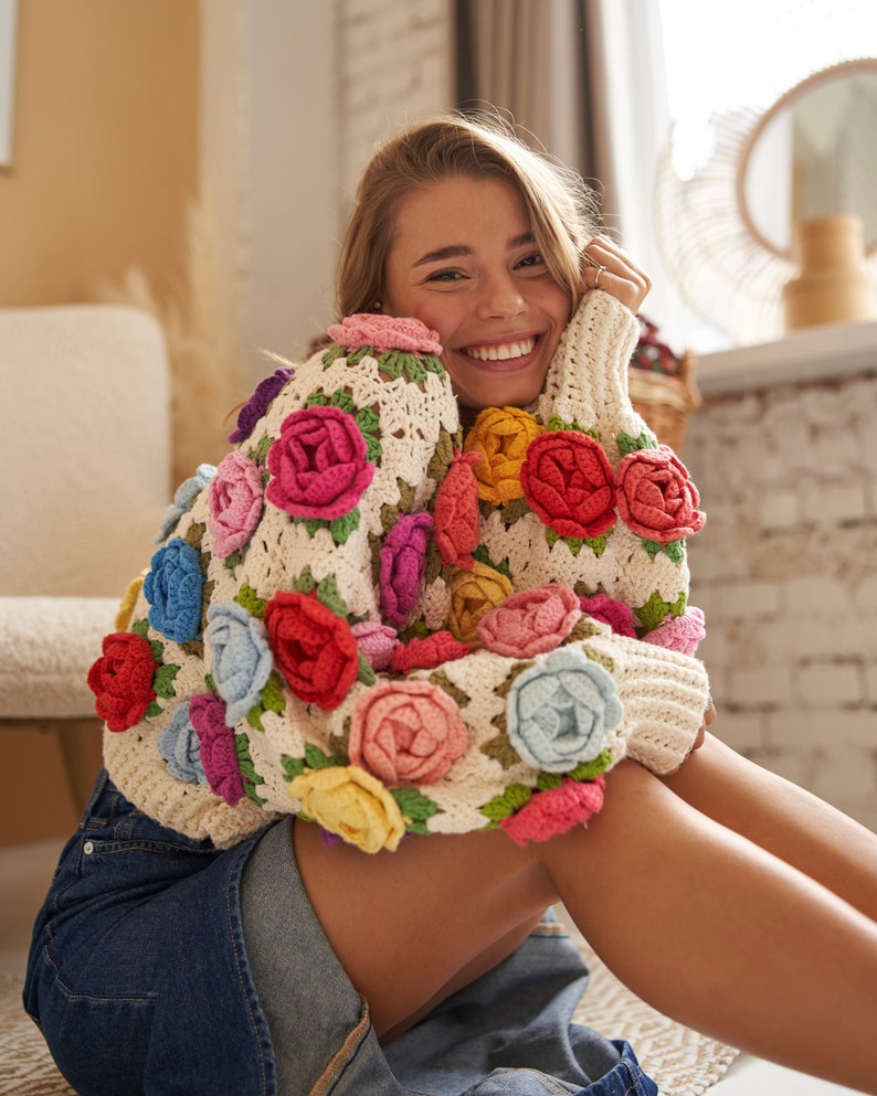 May include: A woman wearing a white crocheted sweater with colorful flowers. The sweater has a granny square pattern and is made with a variety of yarn colors.