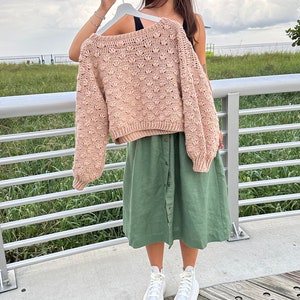 May include: A light pink crocheted sweater with a textured design, worn over a green skirt and white high-top trainers.