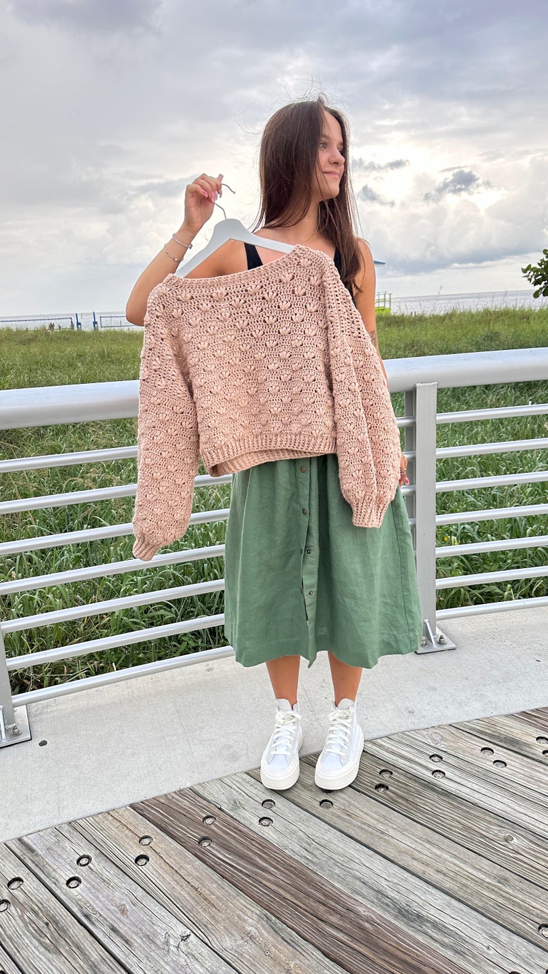 May include: A light pink crocheted sweater with a textured design, worn over a green skirt and white high-top trainers.