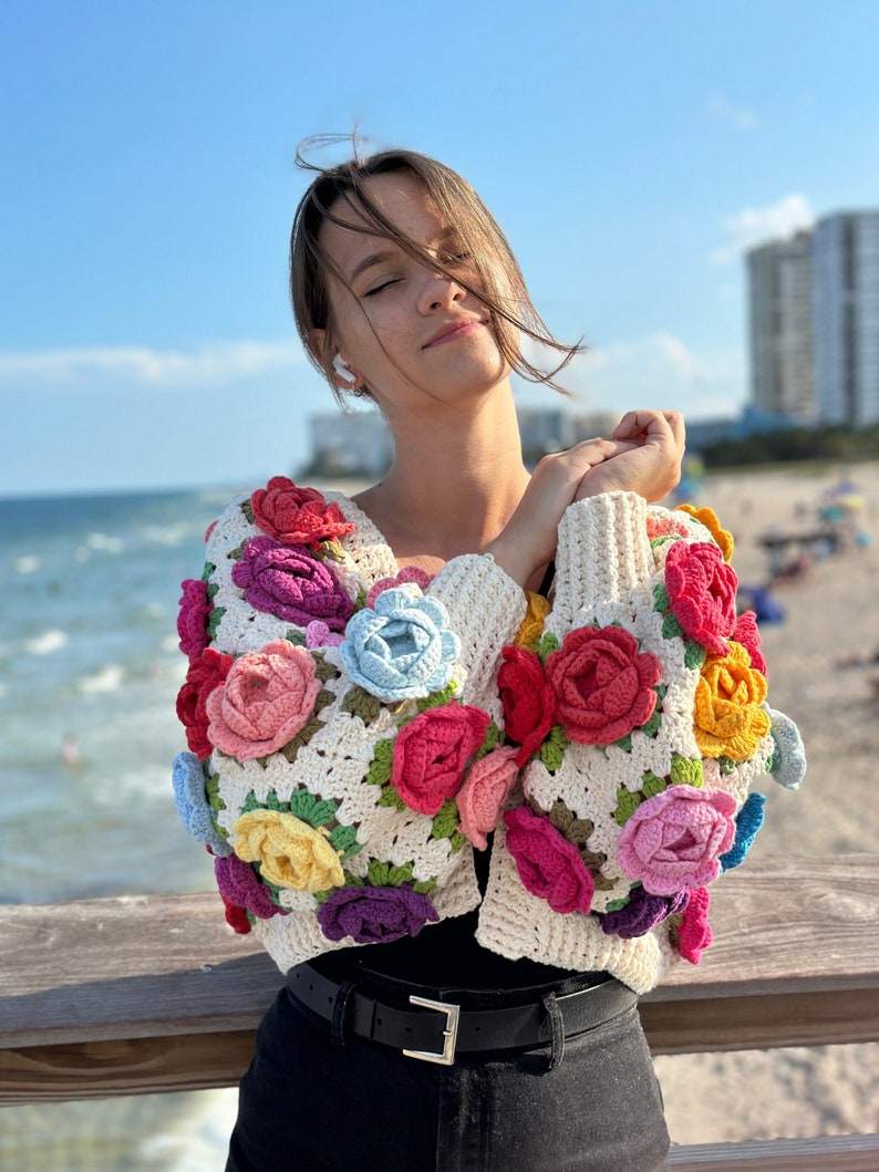 May include: A woman wearing a white crocheted cardigan with colorful flower embellishments. The cardigan has a square stitch pattern and features a variety of colors, including pink, red, yellow, blue, and purple.