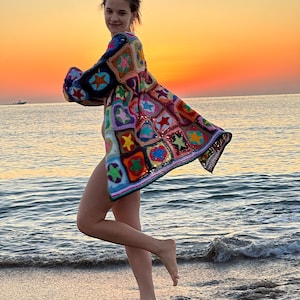 Op de afbeelding: Een vrouw draagt een kleurrijk gehaakt vest met een sterrenpatroon, staand op een zandstrand met de zee op de achtergrond. De lucht is een levendige oranje en roze.