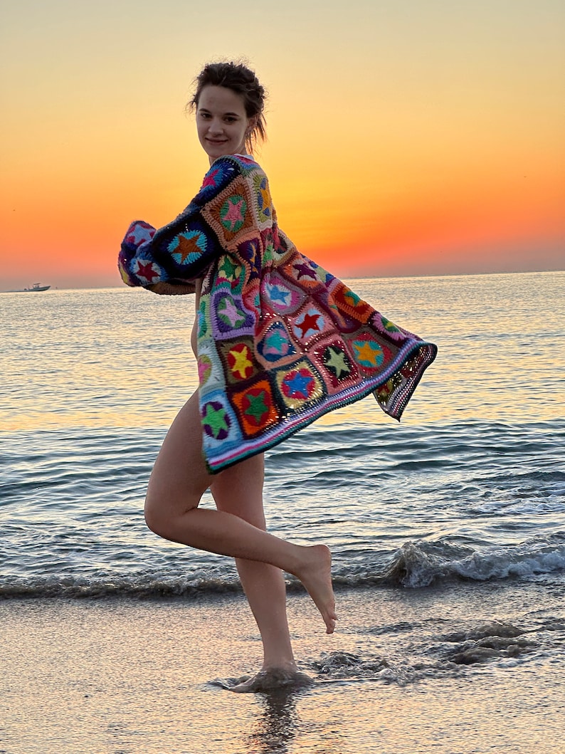 Op de afbeelding: Een vrouw draagt een kleurrijk gehaakt vest met een sterrenpatroon, staand op een zandstrand met de zee op de achtergrond. De lucht is een levendige oranje en roze.