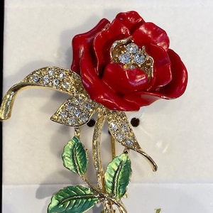 May include: A gold-tone brooch featuring a red enamel rose with clear rhinestone accents. The rose stem is adorned with green enamel leaves. The brooch is labeled "La Scala".