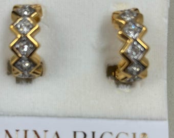 Op de afbeelding: Een paar gouden creolen met een zigzag-ontwerp en heldere stenen. De oorbellen zijn op een witte achtergrond met de tekst "NINA RICCI" eronder.