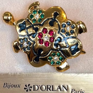 Op de afbeelding: Goudkleurige schildpadbroche met bloemmotieven. De broche heeft rode, groene en blauwe edelstenen, met een centrale cluster van rode stenen. Van Bijoux D'Orlan Paris.