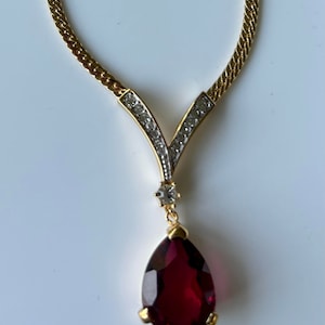 Op de afbeelding: Een gouden ketting met een traanvormige rode edelsteen hanger. De hanger is versierd met kleine heldere strass steentjes.