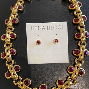 Könnte beinhalten: Eine goldene Halskette mit roten Kristall-Akzenten und passenden Ohrringen. Der Schmuck wird auf einer weißen Karte mit dem aufgedruckten Text "NINA RICCI" präsentiert.
