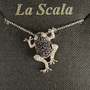 May include: A silver necklace with a black and white frog pendant. The pendant is made of sterling silver and is adorned with black and white cubic zirconia stones. The necklace is displayed on a black background with the words "Bijoux La Scala" and "Sterling Silver".