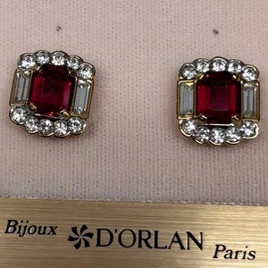 May include: Pair of gold-toned clip-on earrings. Each earring features a large, rectangular, deep red gemstone surrounded by clear, round and baguette-cut crystals. The earrings are set against a pink background, with the text "Bijoux D'ORLAN Paris" visible.