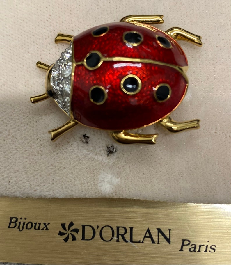 May include: A gold ladybug brooch with red enamel and black spots. The ladybug has a row of clear rhinestones on its back. The brooch is on a beige surface with the words 'Bijoux D'ORLAN Paris' visible below.
