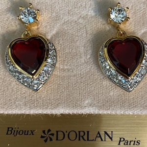 May include: A pair of gold heart-shaped earrings with red glass centres and clear crystal accents. The earrings are labelled "Bijoux D'ORLAN Paris".