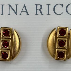 May include: A pair of gold tone earrings with three red stones each. The earrings are round and have a geometric design.