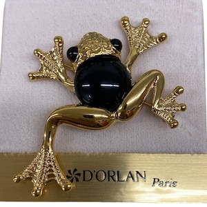 May include: A gold-toned frog brooch with a large black center and black eyes. The brooch is detailed with textured skin and outstretched limbs. The brand name "D'ORLAN Paris" is printed on the bottom.