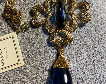 Puede incluir: Un collar de oro con un colgante de cristal azul. El colgante tiene forma de lágrima y presenta un diseño delicado e intrincado. El collar está etiquetado como "Nina Ricci Paris".