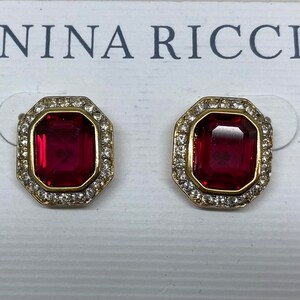 Puede incluir: Un par de pendientes de oro con detalles de piedras preciosas rojas y un halo de cristal transparente. Los pendientes están etiquetados con "NINA RICCI".