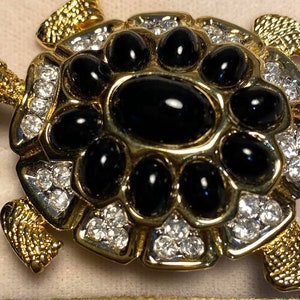 May include: A gold-toned turtle brooch featuring black onyx stones and clear rhinestones. The turtle's shell is adorned with a large black onyx stone surrounded by smaller black onyx stones and rhinestones.