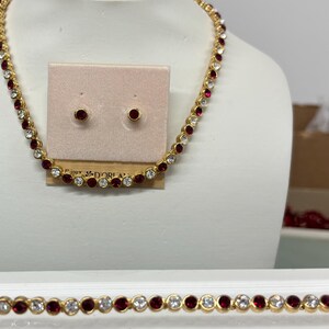 Könnte beinhalten: Ein goldfarbenes Schmuckset mit Halskette, Ohrringen und Armband. Halskette und Armband sind mit abwechselnd roten und klaren Edelsteinen verziert. Die Ohrringe sind kleine, runde, rote Edelsteine. Der Schmuck wird auf einer weißen Schaufensterpuppe präsentiert.
