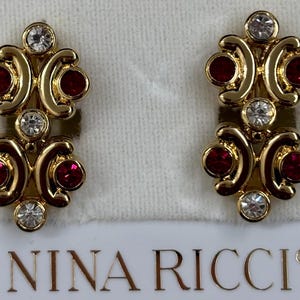 May include: Gold tone earrings with red and clear crystals. The earrings are designed with a double-loop shape and feature the text "NINA RICCI" on the card.