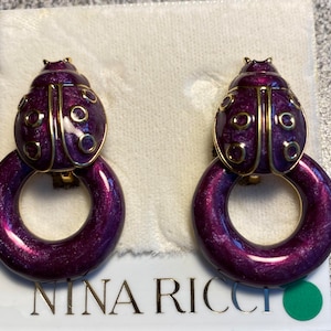 Puede incluir: Un par de pendientes de color morado y dorado con forma de mariquita y diseño de anillo. Los pendientes están sobre un fondo blanco con el texto "NINA RICCI" visible.