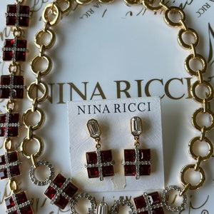 May include: A gold chain necklace with red and clear crystal accents. The necklace is displayed with matching earrings with red and clear crystal accents. The packaging includes the brand name "Nina Ricci".