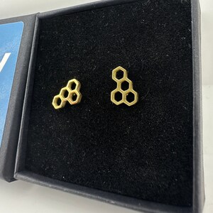Könnte beinhalten: Ein Paar goldfarbene Honigwaben-Ohrstecker, präsentiert in einer schwarzen Schmuckschatulle. Die Ohrringe sind klein, geometrisch und weisen ein sechseckiges Design auf. Die Schatulle ist geöffnet und zeigt die Ohrringe auf dunklem Samt.