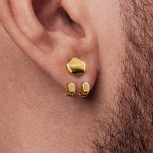 May include: Close-up of an ear with two gold earrings. The top earring is an irregular shape, and the lower earring has two small, rectangular shapes. The person has a beard.