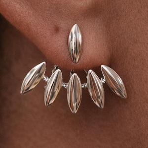 May include: Silver-toned earrings with a unique design. The earrings feature a small, elongated stud and a fan-like arrangement of six similar, curved elements. The jewelry is worn on a person's ear, with the skin tone providing a contrasting backdrop.