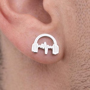 Peut inclure: Boucle d'oreille en argent en forme de casque avec un motif d'onde sonore. La boucle d'oreille est portée sur l'oreille d'une personne. Le design est une représentation minimaliste de la musique.